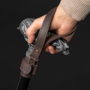 May include: A brown leather strap with a metal buckle is attached to a black walking stick. The stick has a silver animal head at the top.