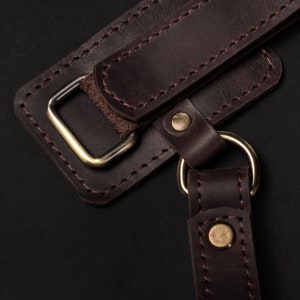 May include: Close-up of a brown leather strap with a metal buckle and stitching. The strap is attached to a ring, which is also made of metal.