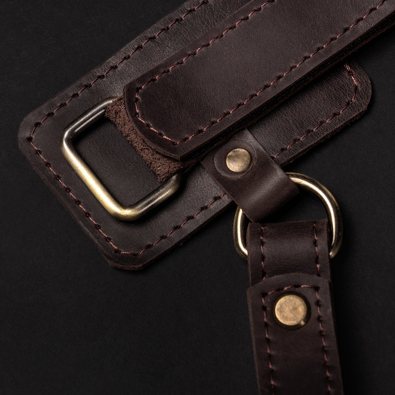 May include: Close-up of a brown leather strap with a metal buckle and stitching. The strap is attached to a ring, which is also made of metal.