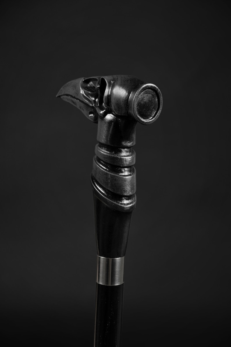 Pode incluir: Uma bengala preta com uma pega detalhada em forma de cabe&ccedil;a de corvo. A pega tem uma banda prateada em torno da base.