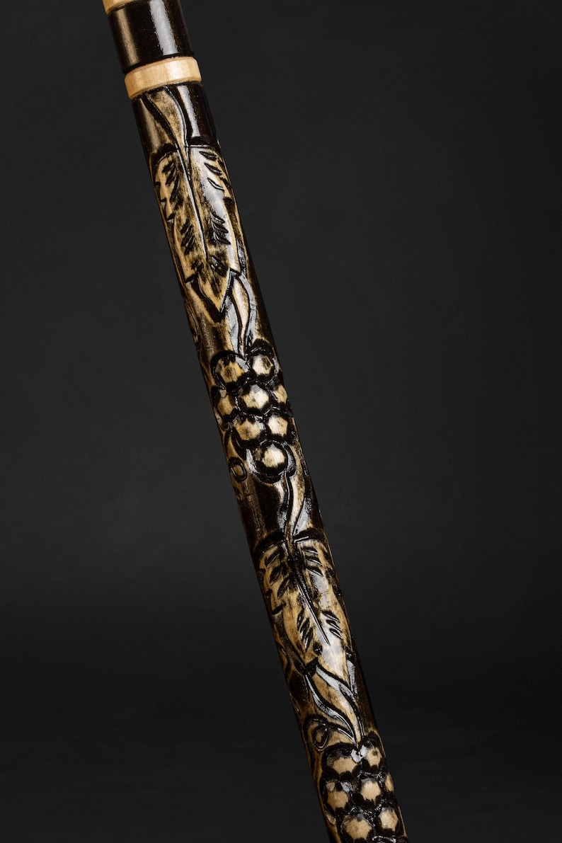 May include: A wooden walking stick with a dark brown finish and intricate carvings of leaves and grapes. The carvings are in a lighter colour, creating a contrast against the dark wood.