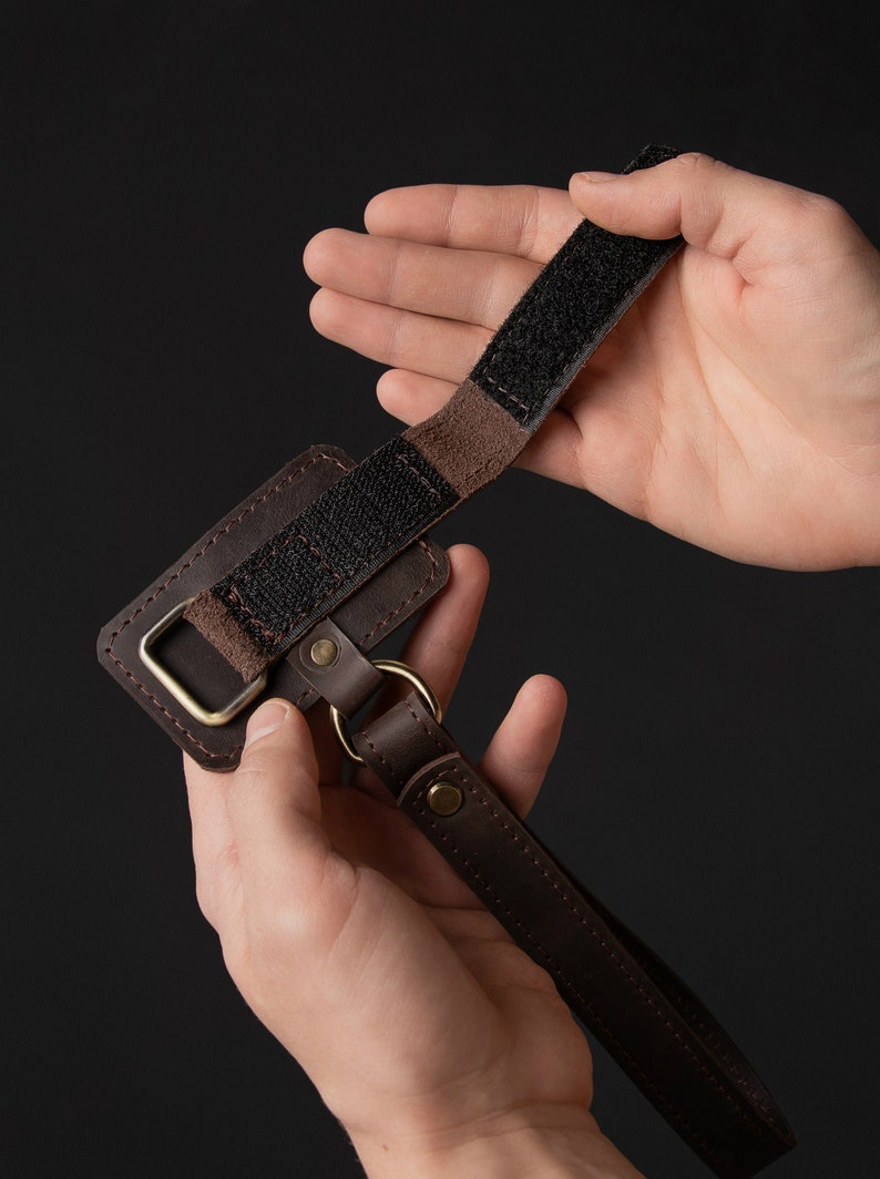 May include: A brown leather camera strap with a black velcro strap and a gold metal ring. The strap is held in two hands.