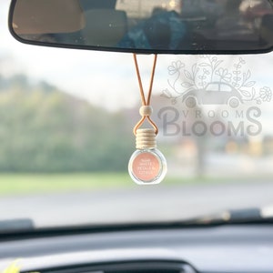 May include: A car air freshener hanging from the rearview mirror. The air freshener is a small glass bottle with a wooden cap and a brown cord. The bottle is filled with a light pink liquid and has a label that reads "Vroom Blooms White Petals & Citrus".