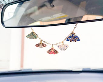 May include: A gold chain with five colourful moth charms hanging from a rearview mirror. The moths are painted in shades of blue, green, pink, white, and black.