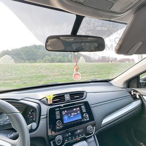 May include: A car interior with a rearview mirror, a small potted plant, and a car air freshener hanging from the mirror. The air freshener is a small glass bottle with a cork stopper and a brown and white string.