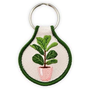 May include: A teardrop-shaped keychain featuring an embroidered design of a green potted plant with a brown stem in a light pink pot. The embroidery is set against a cream-colored fabric background with a dark green stitched border and a metal ring.
