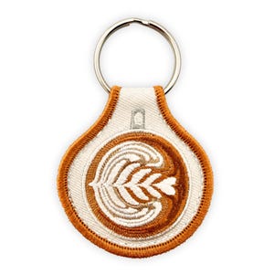 Puede incluir: Un llavero bordado redondo con un diseño de arte latte de café en tonos marrón y blanco. El parche de tela circular tiene un borde naranja grueso y cosido, y está unido a una anilla de metal plateado para las llaves.