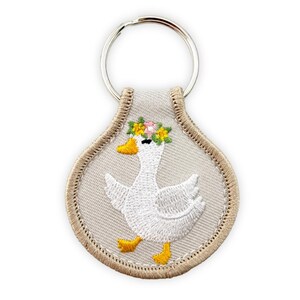 May include: A teardrop-shaped keychain featuring an embroidered white goose with a yellow beak and feet. The goose wears a colorful floral crown of pink and yellow flowers. The background is light beige with a matching beige stitched border.