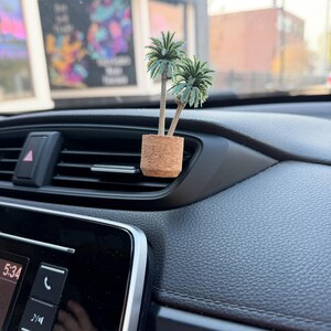 May include: A miniature palm tree air freshener. The small, artificial palm trees are in a cork pot, designed to clip onto a car's air vent. The car's dashboard is black.