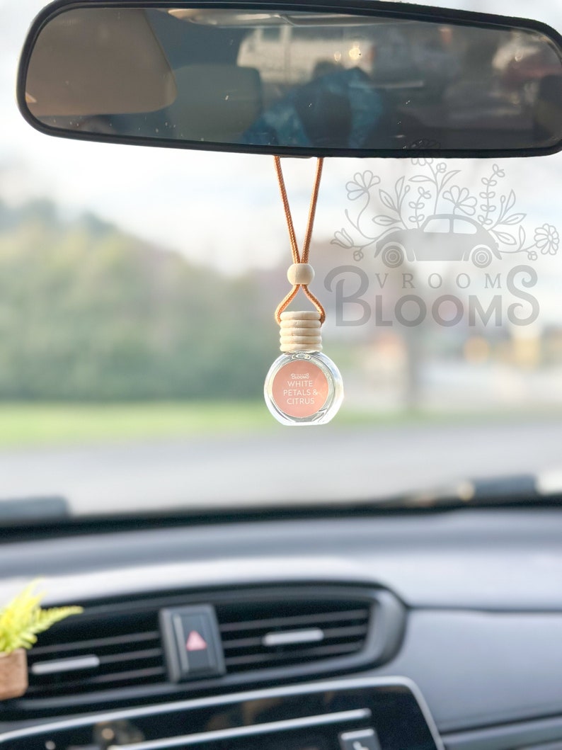 May include: A car air freshener hanging from the rearview mirror. The air freshener is a small glass bottle with a wooden cap and a brown cord. The bottle is filled with a light pink liquid and has a label that reads "Vroom Blooms White Petals & Citrus".