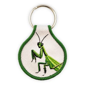 May include: A teardrop-shaped keychain featuring an embroidered green praying mantis on a white fabric background. The design is bordered by a thick green stitched edge and is attached to a circular silver-toned metal split ring for keys.