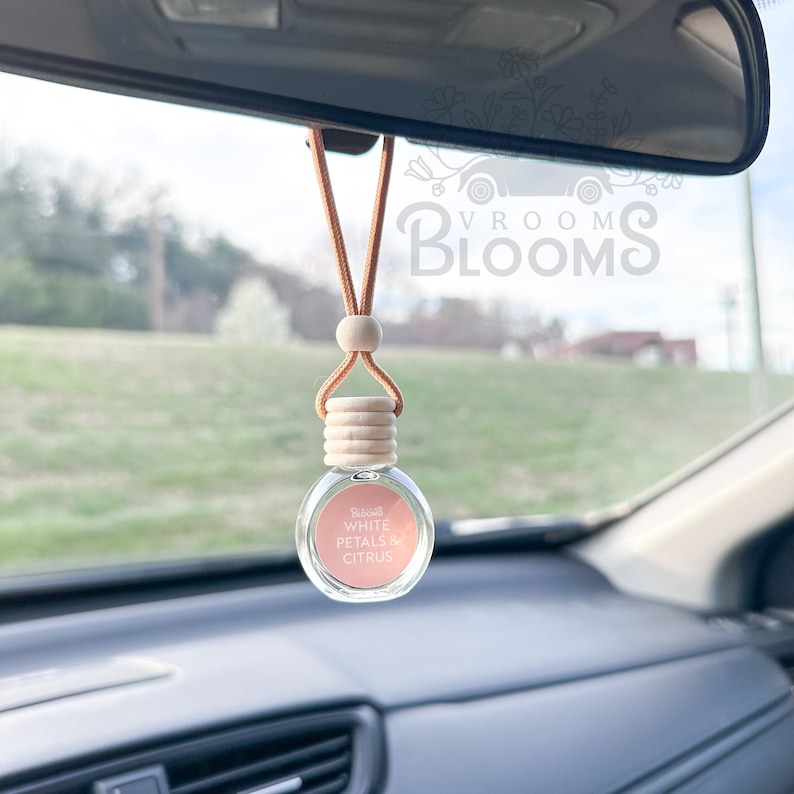May include: A brown leather car air freshener with a wooden bead and a clear glass bottle. The bottle has a white label with the text "Bloom" and "White Petals & Citrus".