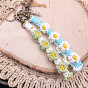 Könnte beinhalten: Zwei gehäkelte Schlüsselanhänger mit Gänseblümchen-Blütenmotiven. Ein Schlüsselanhänger ist weiß mit hellblauen Akzenten und der andere ist weiß mit grünen Akzenten. Beide Schlüsselanhänger haben gelbe Zentren auf den Blüten.