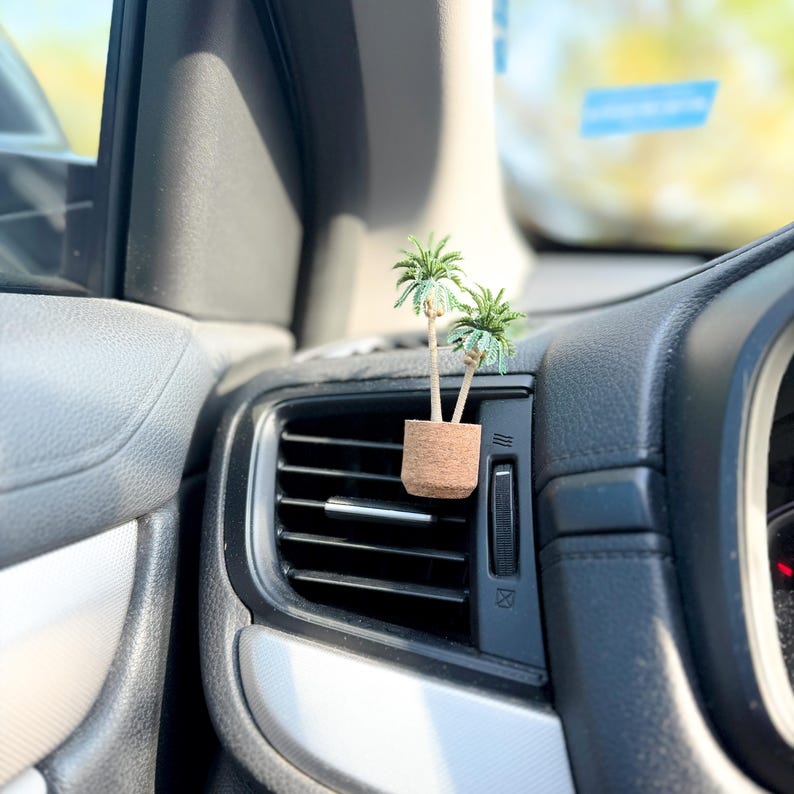 May include: A miniature palm tree air freshener in a cork pot, clipped to a car's air vent. The small, green palm trees have detailed fronds, and the pot is a natural brown color. The car's interior is black and gray.