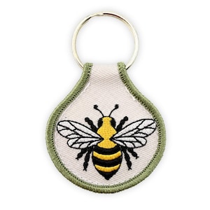May include: A teardrop-shaped embroidered keychain featuring a central yellow and black bee design on a cream-colored fabric background. The keychain is bordered by a thick olive green stitched edge and includes a circular silver metal ring.