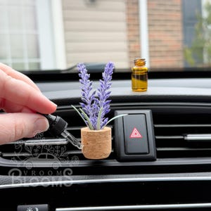 Puede incluir: Un ambientador de coche con forma de planta de lavanda. La planta está en una pequeña maceta de corcho y está unida a una ventilación de coche. También se puede ver una pequeña botella de aceite esencial en la imagen.