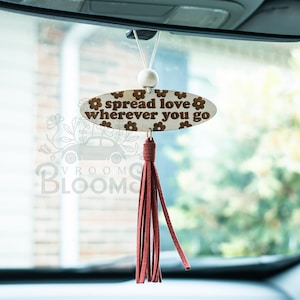 May include: A wooden oval car charm with the text "spread love wherever you go" and a brown floral pattern. The charm is hanging from a white string with a white bead and a red tassel.