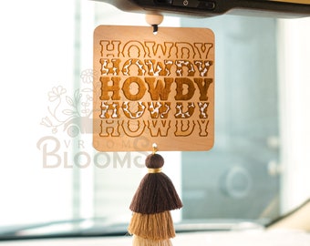May include: Wooden car air freshener with a brown tassel. The air freshener is rectangular and has the word "Howdy" written on it multiple times in a cow print pattern.