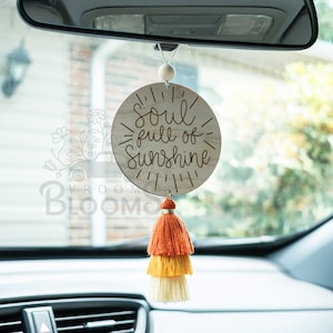 May include: A wooden car charm with the text "Soul full of Sunshine" and a multi-colored tassel hanging from it. The charm is hanging from the rearview mirror of a car.