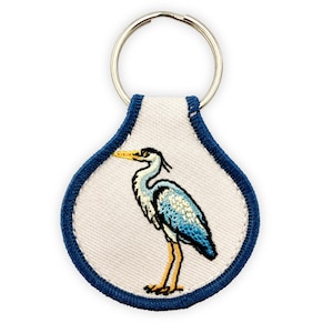 May include: A teardrop-shaped keyring featuring an embroidered grey and blue heron with a yellow beak on a cream-coloured fabric background. The design is bordered by a thick navy blue stitched edge and includes a silver metal split ring at the top.