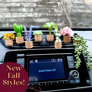 Può includere: Una collezione di piante artificiali in miniatura in piccoli vasi di sughero fissati alla bocchetta dell'aria di un'auto. Le piante includono un girasole, un cactus verde, lavanda viola, una succulenta, una pianta verde, fiori bianchi, fiori rosa e una pianta rampicante.