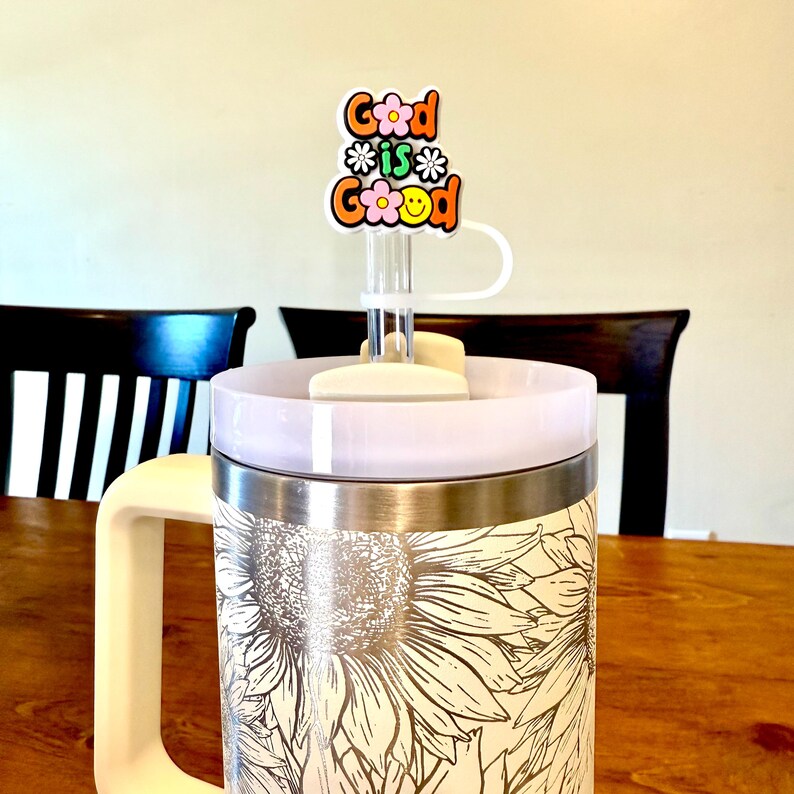 Puede incluir: Una taza de viaje de acero inoxidable blanca con un dise&ntilde;o de girasol. La taza tiene una tapa blanca con una pajita de pl&aacute;stico transparente y un adorno de pajita colorido que dice "Good is Good" con flores y una cara sonriente.