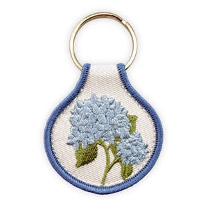 May include: A teardrop-shaped keychain featuring an embroidered design of light blue hydrangea flowers with green leaves on a white fabric background. The edges are finished with a blue stitched border and a gold-toned metal split ring.