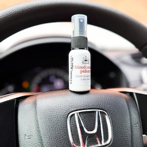 May include: A white car air freshener spray bottle with black accents and a clear spray nozzle sits on a black steering wheel. The bottle has the text "Vroom Spray" and "Blood on Palomino" with "Odor Eliminator Car Spray".