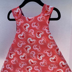 May include: A sleeveless red dress with a white heart pattern. The dress has a scoop neck and two buttons on each shoulder strap.