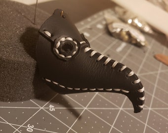 May include: A black leather plague doctor mask with white stitching and a round eye piece. The mask is on a gray cutting mat.