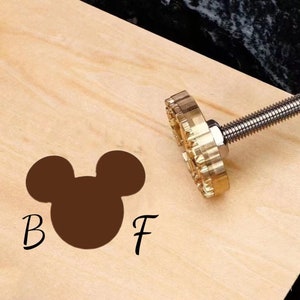 以下が含まれることがあります： 木製の表面にミッキーマウスの頭のデザインと「B」と「F」の文字が刻印された真鍮製のスタンプです。