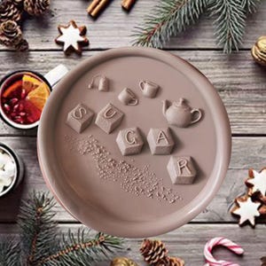 May include: A round, light brown silicone mold featuring embossed designs of a teapot, a teacup, a tea bag, and five square blocks spelling out the word SUGAR. The surface of the mold includes small, scattered circular indentations.