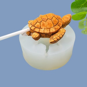 Op de afbeelding: Een witte siliconen mal voor het maken van een kleine cakepop in de vorm van een schildpad. De mal heeft een gat in het midden om een ​​lollipopstokje in te steken. Een bruine cakepop in de vorm van een schildpad zit in de mal.