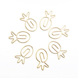 May include: A collection of gold-coloured metal paper clips, each shaped like a pineapple, arranged in a circle on a white backdrop. The clips have a looped body and a detailed crown.