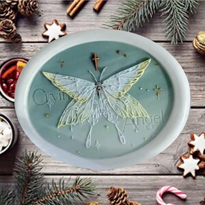 Pode incluir: Um prato decorativo ovalado azul claro com uma borda branca. O prato apresenta um design detalhado de borboleta com asas brancas e amarelas, uma cruz e as palavras "Crying Angel". O prato é rodeado por decorações de Natal.