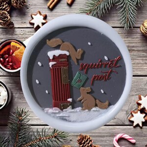May include: A round, gray decorative plate with a red post box and two squirrels. The plate has the words "squirrel post" in red script. The plate is surrounded by a white border and is decorated with snow.