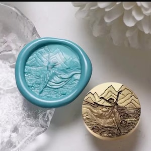 Puede incluir: Un sello de cera turquesa con un diseño en relieve de una mujer bailando frente a montañas y un lago. El sello de cera está sobre una superficie de vidrio transparente. Se muestra un segundo sello de cera con un diseño similar en oro.