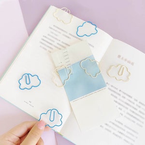 Op de afbeelding: Een set van zes wolkvormige paperclips in twee kleuren: blauw en wit. De clips zijn gerangschikt op een wit notitieboek met een blauw-witte pagina.