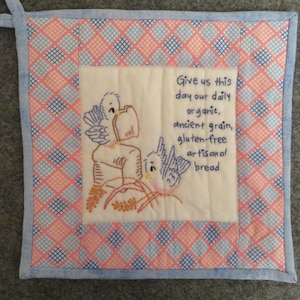 May include: Square quilted potholder with a blue and pink diamond pattern border. The center features embroidered birds and text that reads "Give us this day our daily organic, ancient grain, gluten-free artisanal bread."