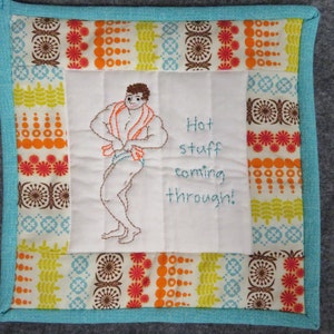 May include: Square quilted pot holder with a turquoise border and hanging loop. The center features an embroidered illustration of a muscular figure with a towel, and the text "Hot stuff coming through!" The border has a colorful retro floral pattern.