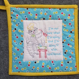 May include: A square quilted potholder with a yellow border and a light blue fabric center featuring a colorful diamond pattern. The white center panel displays an embroidered sheep wearing a chef hat, stirring a bowl, with the text: I'm not sure what I'm doing, but I'm doing it with confidence!