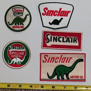 May include: A selection of vintage Sinclair Oil patches featuring the Sinclair Dino dinosaur logo. The patches are in various shapes, including circles and rectangles, with text such as "GASOLINE" and "MOTOR OIL". The patches are green, red, and white.