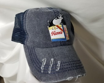 May include: A blue denim trucker hat with a black mesh back. The hat has a distressed look and features an embroidered patch of a cartoon bear sitting on a box of Hamm's beer.
