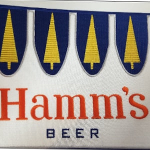 May include: Embroidered patch featuring the Hamm's Beer logo. The design includes a white background with the brand name in orange, and the word "BEER" in blue. Above the text are four blue crests with yellow tree designs.