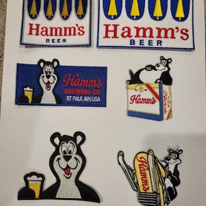 May include: Five vintage Hamm's Beer patches. Two patches feature the Hamm's logo with a row of pine trees. One patch features a bear holding a beer mug. One patch features a bear sitting on a Hamm's beer can. One patch features a bear riding a Hamm's beer can.