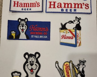May include: Five vintage Hamm's Beer patches. Two patches feature the Hamm's logo with a row of pine trees. One patch features a bear holding a beer mug. One patch features a bear sitting on a Hamm's beer can. One patch features a bear riding a Hamm's beer can.