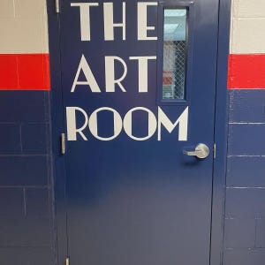 May include: A dark blue metal door featuring the white text THE ART ROOM in a bold, sans-serif font. The door includes a rectangular window with a wire-reinforced glass pane and a silver-colored horizontal lever handle on the right side.