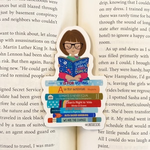 May include: A stack of colorful books with feminist and political messages. The books are titled "Good Night Rebel Girls", "A is for Activist", "Separate is Never Equal", "Lilian's Right to Vote", "Alter Masters: Women's Right to Vote", "Ruth Bader Ginsburg", and "We Are the Change". The books are stacked on top of each other and are held together by a sticker that says "V is for Voting".