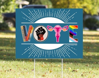 May include: A blue yard sign with the word "VOTE" spelled out in large white letters. Each letter is decorated with a different symbol: a stack of books, a raised fist, a uterus, and a rainbow pride flag. The sign has a white sunburst design behind the letters.
