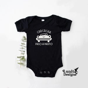 以下が含まれることがあります： 白い文字で「Carcacha Paso a Pasito」と書いてあり、ヴィンテージカーの白いシルエットが描かれた黒いベビーロンパースです。